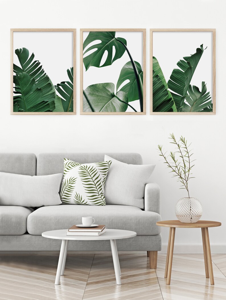 May include: Three framed prints of tropical leaves in shades of green against a white background. The prints are arranged in a row on a white wall.