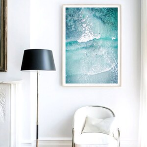 May include: A framed photograph of an aerial view of turquoise ocean water with white waves crashing. The photo is hanging on a white wall in a room with a white chair and a black floor lamp.
