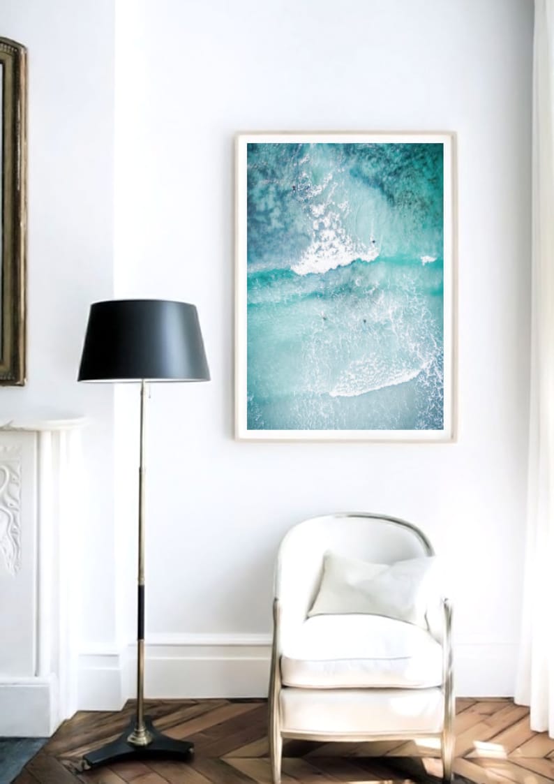 May include: A framed photograph of an aerial view of turquoise ocean water with white waves crashing. The photo is hanging on a white wall in a room with a white chair and a black floor lamp.