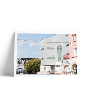 May include: A white building with a sign that says "VENICE" in large, illuminated letters. The building is pink and white and has a blue archway. The sign is hanging from a wire above the building.