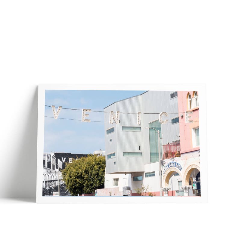 May include: A white building with a sign that says "VENICE" in large, illuminated letters. The building is pink and white and has a blue archway. The sign is hanging from a wire above the building.