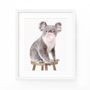 Op de afbeelding: Een ingelijste print met een grijze koala die op een kleine houten kruk zit. De koala heeft een roze kauwgomballenbel in zijn mond. De print staat tegen een witte achtergrond.
