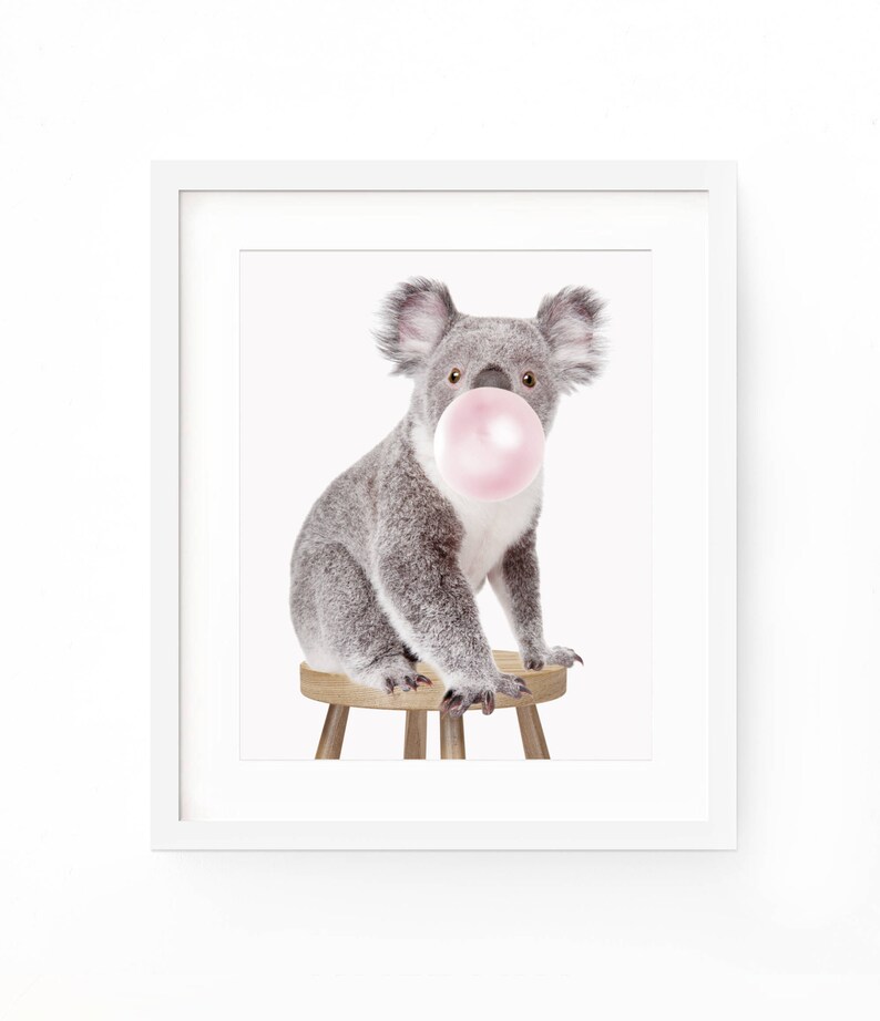 Op de afbeelding: Een ingelijste print met een grijze koala die op een kleine houten kruk zit. De koala heeft een roze kauwgomballenbel in zijn mond. De print staat tegen een witte achtergrond.