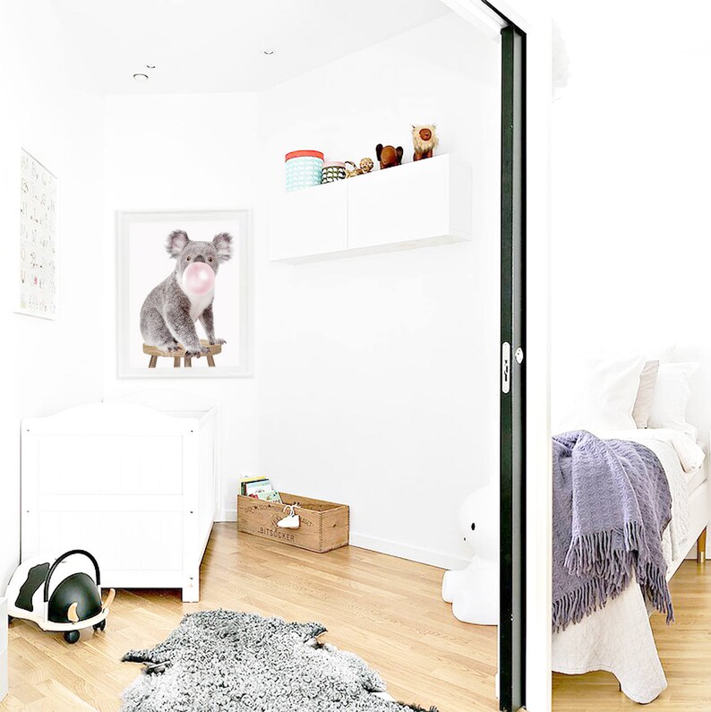 Op de afbeelding: Een heldere, witte kinderkamer met een ingelijste koala-print die een kauwgomblaas blaast. Een witte wieg, een houten speelgoedkist en een zwart-wit speelgoed om op te rijden zijn zichtbaar. Een grijs tapijt en een bed met een paarse deken zijn ook in de kamer.