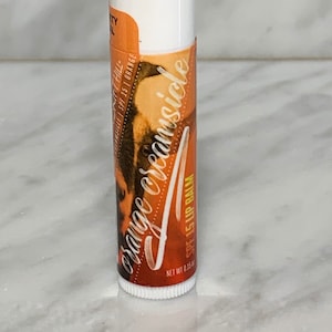 May include: A white and orange lip balm tube with the words "Orange creamsicle" in a cursive font. The tube also has the text "SPF 15 Lip Balm" and "Net wt 0.15 oz". The background is a marble pattern.