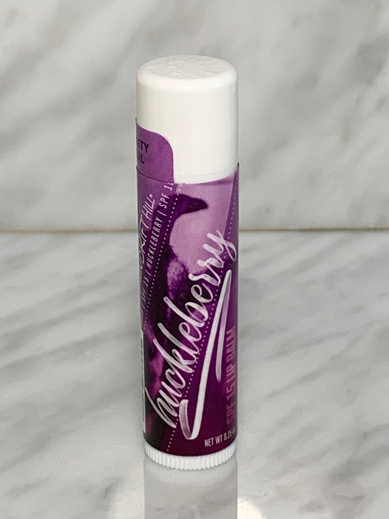 May include: A purple lip balm tube with a white cap. The label says "Huckleberry" and "SPF 15 Lip Balm".