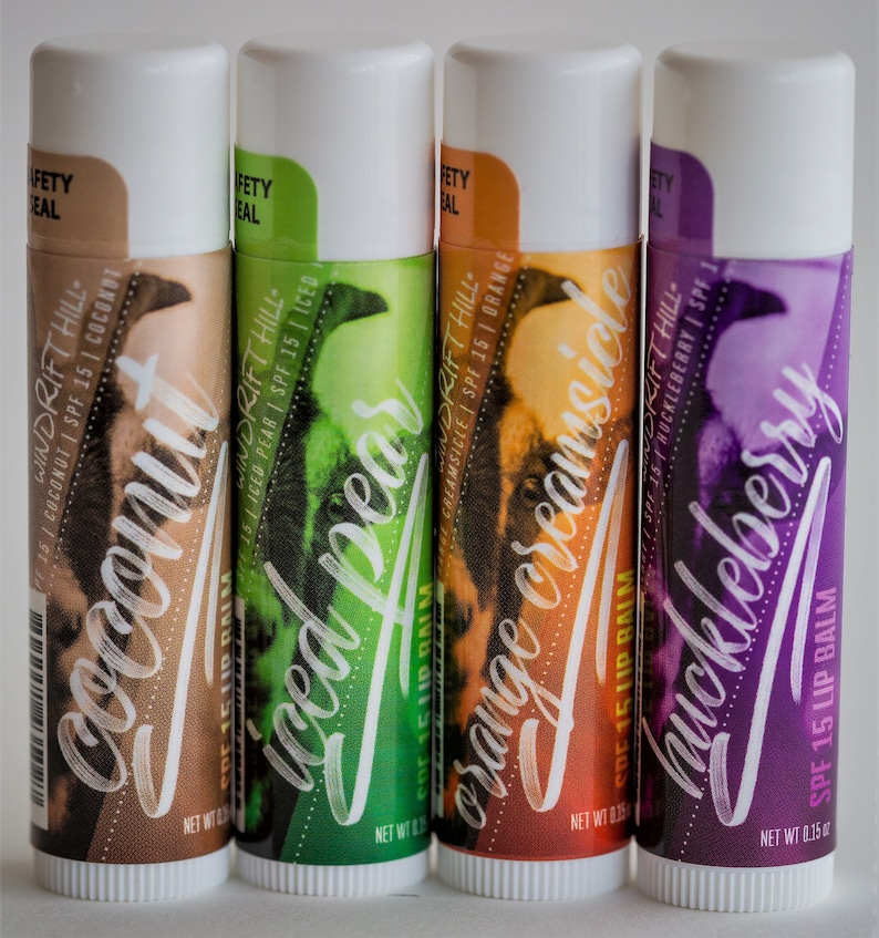 May include: Four lip balm tubes with different flavors: Coconut, Iced Pear, Orange Creamsicle, and Huckleberry. Each tube has a white cap and a colorful label with the flavor name and a graphic design. The lip balms are SPF 15.