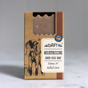 May include: A brown cardboard box with a white label that says "WINDRIFT HILL MOISTURIZING GOATS MILK SOAP Honey N' Rolled Oats". The box has a picture of a goat on it and a circle with the text "MADE IN MONTANA USA".