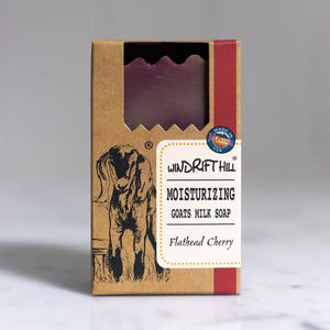 May include: A brown cardboard box with a red stripe and a goat illustration. The box contains a bar of purple goat milk soap with the text "Windrift Hill Moisturizing Goats Milk Soap Flathead Cherry" printed on the front.