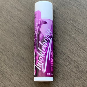 May include: A white tube of huckleberry flavored lip balm with SPF 15. The tube has a purple and white design with the words "WINDRIFT HILL" and "huckleberry" written on it.