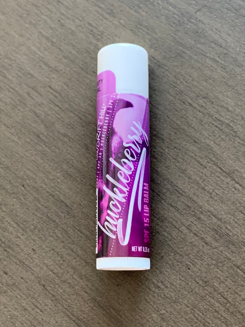 May include: A white tube of huckleberry flavored lip balm with SPF 15. The tube has a purple and white design with the words "WINDRIFT HILL" and "huckleberry" written on it.
