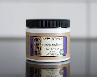Könnte beinhalten: Ein weißes Glas mit schwarzem Deckel und einem braunen Etikett. Das Etikett lautet "BODY BUTTER Montana Huckleberry Very Dry Skin 4 oz. With Goat Milk, Shea & Mango Butters".