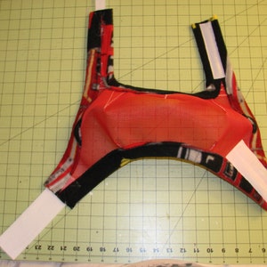 Puede incluir: Un arn&eacute;s para perro de tela roja y negra con correas de velcro blancas. El arn&eacute;s tiene un forro de malla.