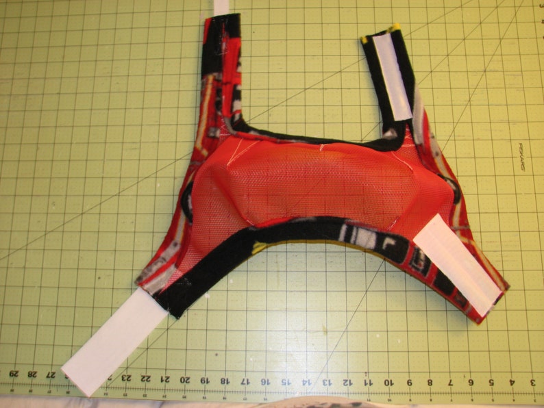 Puede incluir: Un arn&eacute;s para perro de tela roja y negra con correas de velcro blancas. El arn&eacute;s tiene un forro de malla.