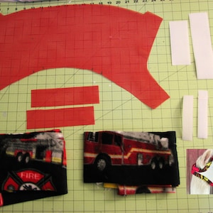 May include: A red fabric pattern with two strips of red fabric, two strips of white fabric, and a piece of black fabric with a fire truck and the word "FIRE" printed on it. A white llama wearing a red and white fabric mask is in the bottom right corner.