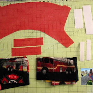 Puede incluir: Un patr&oacute;n de tela roja con dos tiras de tela roja y cuatro tiras de tela blanca. Hay dos piezas de tela con un estampado de cami&oacute;n de bomberos y logotipo del departamento de bomberos. Una pieza de tela est&aacute; doblada y la otra est&aacute; enrollada. Hay una foto de un caballo con una m&aacute;scara de tela roja y negra.