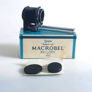 May include: A black Spiratone Macrobel bellows camera extension for Nikon cameras, in its original box with two lens caps. The box is blue and white with the text "Spiratone Rapid-rail Macrobel Bellows for Nikon".