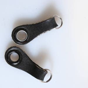 Peut inclure: Deux porte-clés en cuir noir avec des anneaux en métal argenté. Chaque porte-clés a un œillet rond en métal.