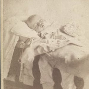 May include: A black and white photograph of a baby sleeping in a crib. The baby is wearing a white gown and is lying on its back with its arms crossed over its chest.