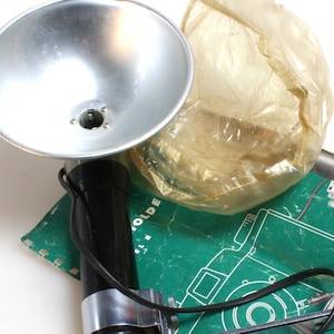 May include: A silver flash unit with a black handle and a clear plastic diffuser. The flash unit is attached to a metal bracket with a screw. The flash unit is connected to a camera by a black wire.