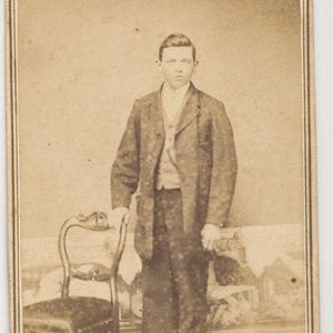 Op de afbeelding: Een zwart-wit portret van een jonge man die voor een stoel staat. Hij draagt ​​een donker colbert en broek. De achtergrond is een wazig beeld van een kamer.