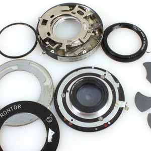 May include: Disassembled parts of a vintage Polaroid camera lens, including the shutter, aperture ring, and lens elements. The lens is labeled "Pronto" and "Polaroid".