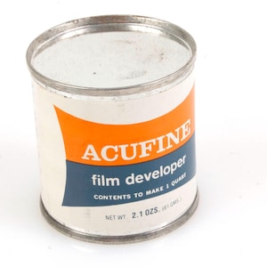 May include: A white metal can with an orange and blue label. The label reads "ACUFINE film developer CONTENTS TO MAKE 1 QUART NET WT. 2.1 OZS. (61 GMS)"