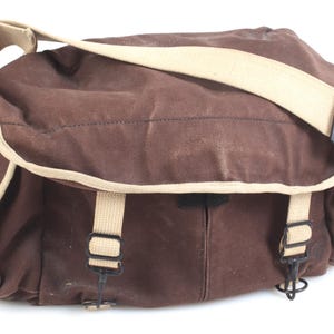May include: A brown canvas messenger bag with a beige shoulder strap and trim. The bag has a rectangular shape with a flap closure and two front straps with buckles. The bag is made of a durable material and is suitable for carrying items.