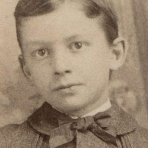 May include: A sepia-toned portrait of a young person, possibly a boy, with short, neatly combed hair. They are wearing a dark suit jacket, a bow tie, and a collared shirt. The image has a vintage aesthetic.