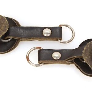 May include: Two dark brown leather keychains with silver key rings and rivets. The keychains feature a unique design with layered, textured accents. The background is plain white.