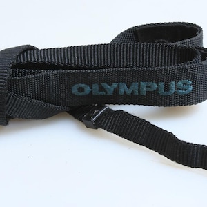 May include: Black Olympus camera strap with the brand name in teal lettering. The strap is made of a woven material and has a plastic clip. The strap is folded over itself.