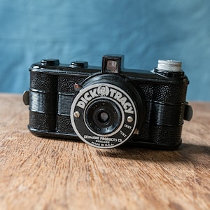 Puede incluir: Una cámara negra Dick Tracy Inst. Time con una lente plateada y una inscripción blanca que dice "Dick Tracy Inst. Time Seymour Products Co. Chicago Made in U.S.A."