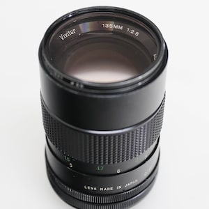 May include: A black Vivitar 135mm f/2.8 lens with a green inscription that reads "Lens Made in Japan".