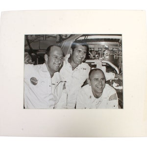 May include: Black and white photograph of three astronauts in their spacesuits, smiling and posing for a photo. The astronauts are standing in front of a spacecraft.