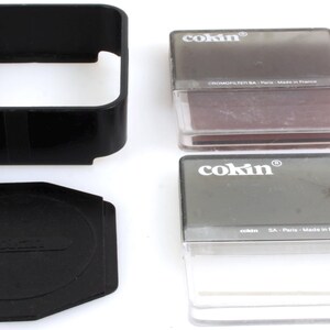 May include: Black Cokin filter holder, lens cap, and two filter cases. The filter cases are clear with grey lids, and the filters inside are brown and grey. The lens cap and holder are black.