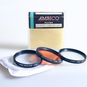 Puede incluir: Un kit de filtros de fotografía con tres filtros con borde negro, un paño de limpieza y un paquete etiquetado como "AMBICO IMAGE SYSTEM FILTER." Los filtros incluyen "HAZE 49mm (UV)," "UW Correction 8706" y "PRO 490 DIFF." Hecho en EE. UU.