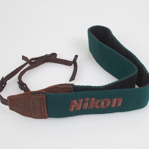 May include: A dark green camera strap featuring the Nikon logo embroidered in brown thread. The strap includes brown leather-like end tabs and thin brown adjustable nylon webbing connectors with black plastic buckles for attaching to a camera body.