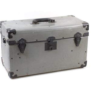 Peut inclure: Une malle de rangement rectangulaire grise dotée d'une poignée en plastique noir sur le couvercle, de renforts d'angle en métal foncé et de trois loquets métalliques frontaux, dont un mécanisme de verrouillage central avec serrure.