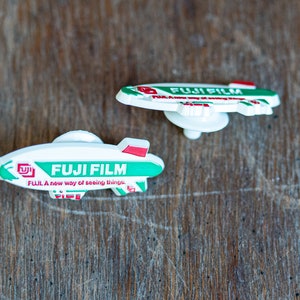 May include: Two white plastic toy blimps with green and red accents. The blimps have the words "FUJI FILM" printed on them. The blimp on the left has the text "FUJI. A new way of seeing things." printed on it.