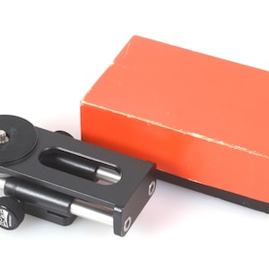 May include: A black metal Novoflex macro focusing rail with a silver adjustment rod and a circular camera mount plate, accompanied by a rectangular orange cardboard box with black base detailing and white text on the side.