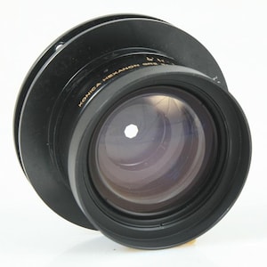 May include: A black Konica Hexanon camera lens with a wide aperture. The lens has a circular design with a series of rings and a clear glass element. The text "KONICA HEXANON" is visible on the lens.