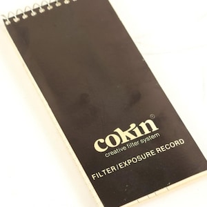 May include: A rectangular spiral-bound notebook with a black cover featuring the white text Cokin creative filter system and FILTER/EXPOSURE RECORD. The notebook has a white edge and a metal wire binding along the top short edge.