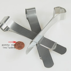 May include: Two silver-colored metal tongs with a U-shaped handle and flat, wide ends. The tongs are arranged in an overlapping cross pattern. A copper-colored penny is positioned to the left with a red arrow pointing to it and the text penny not included.