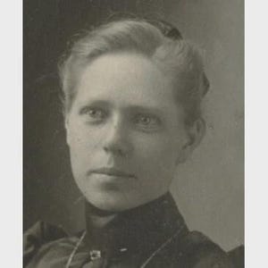 Puede incluir: Un retrato fotográfico vintage en blanco y negro de una persona con ojos claros y cabello peinado en un recogido. El individuo viste una prenda oscura de cuello alto con un detalle de botón visible en el escote, capturado en un primer plano.