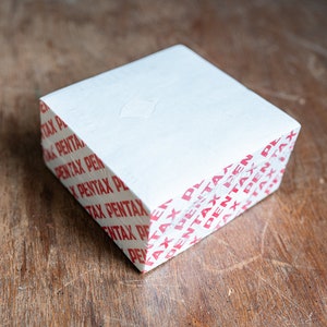 May include: A white box with the word "PENTAX" printed in red on all sides.