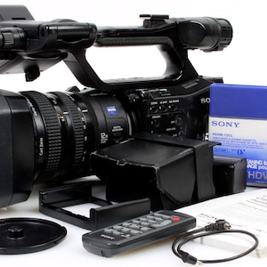 Puede incluir: Una videocámara Sony HDV negra con un objetivo, un mando a distancia, una tapa de objetivo negra, un cargador de batería negro, una cinta Sony HDV azul y un manual.