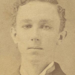 May include: Sepia-toned portrait of a man, likely from the late 19th or early 20th century. The subject has light skin, curly hair, and is wearing a dark suit with a white collared shirt. The image is a head and shoulders shot.