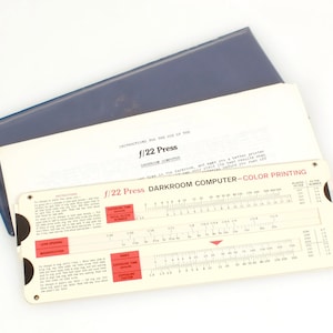 Pode incluir: Uma régua de cálculo branca f/22 Press Darkroom Computer - Color Printing com marcas vermelhas e pretas. A régua de cálculo é usada para calcular os tempos de exposição para impressão a cores.