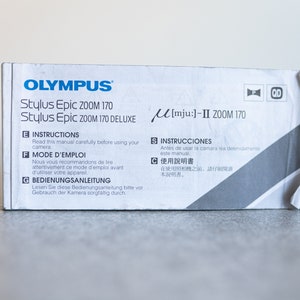 May include: A white box with the text "OLYMPUS Stylus Epic 200M 170 Stylus Epic 200M 170 DELUXE" printed on it. The box also has the text "[mju:]-II ZOOM 170" printed on it. The box has instructions in English, French, German, Spanish, and Japanese.