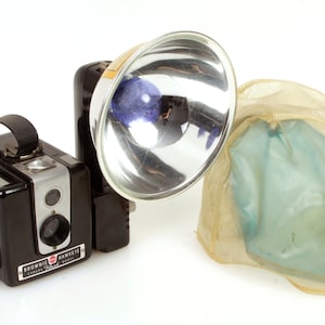 May include: A black Brownie Hawkeye Flash camera with a silver flash attachment and a clear plastic flash cover.